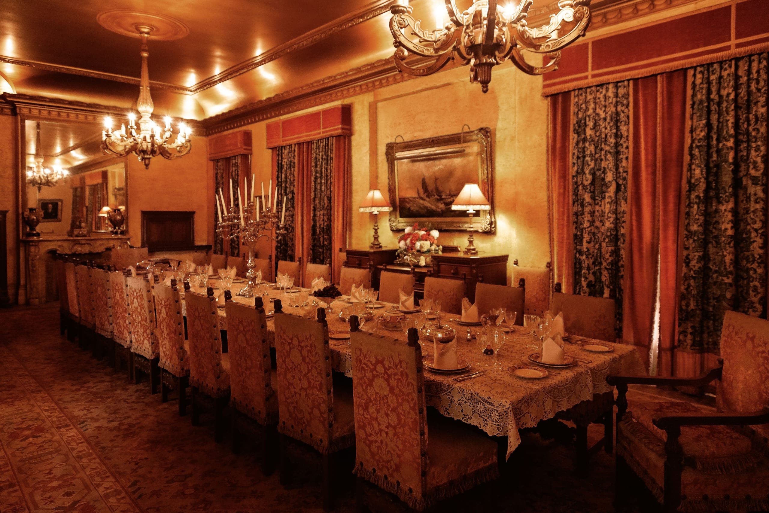 A long candlelit banquet table set with fine china and silver candelabra in a warm gold-lit formal dining room, empty upholstered chairs receding into the distance